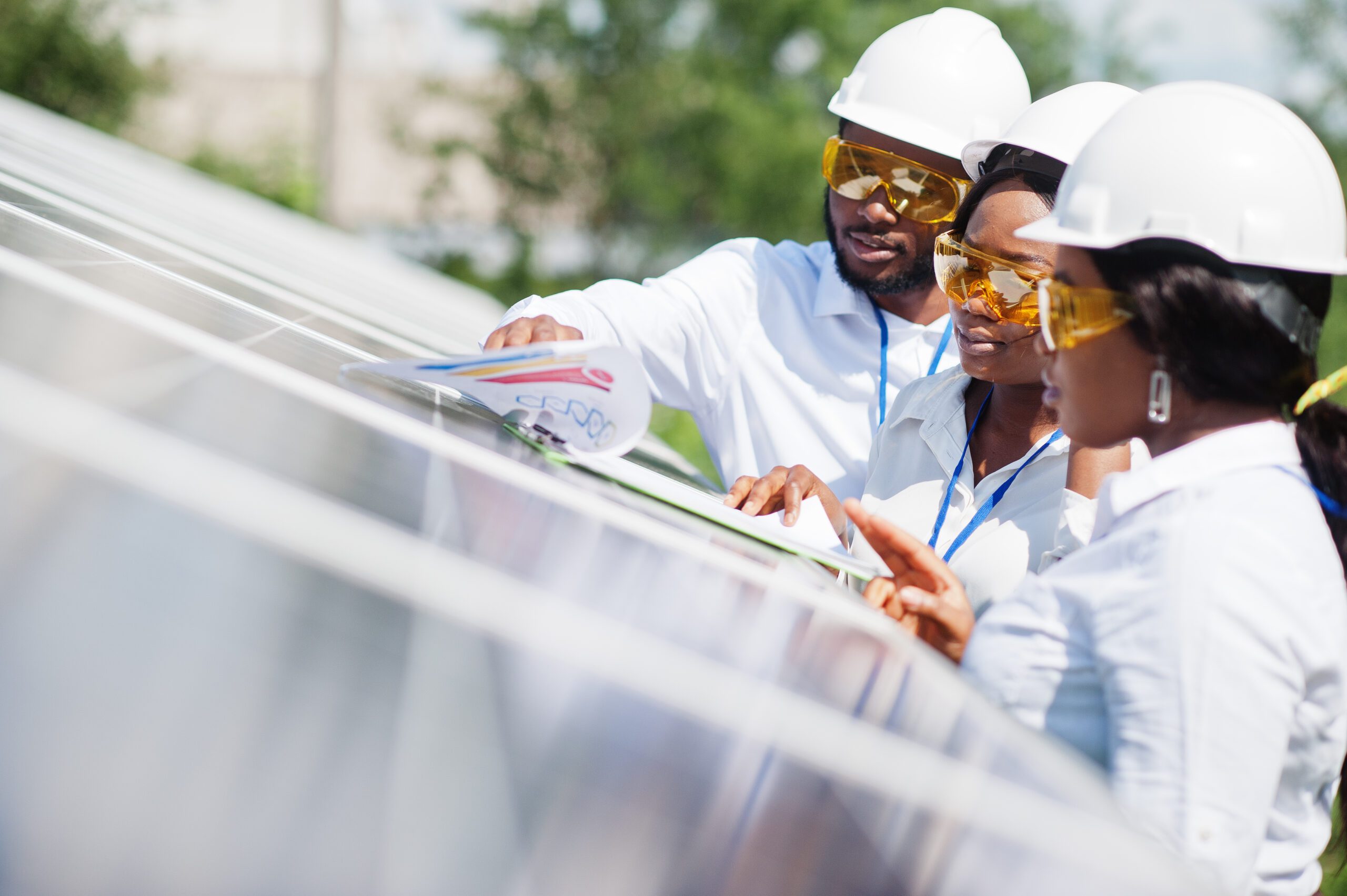 Three people wearing protective equipment standing near a solar panel and looking at some paperwork.